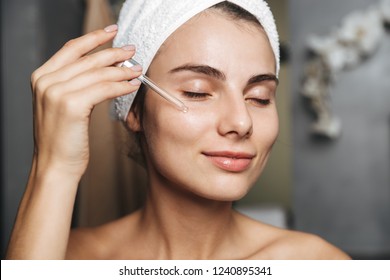 Photo Of Lovely Woman With Towel On Head Applying Cosmetic Oil On Her Face While Standing In Bathroom