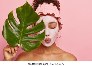 Photo of lovely young Afro American woman applies nourishing clay mask, has eyes shut, lips folded, covers half of face with big green leaf, wears shower headband, isolated on pink background