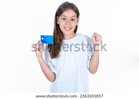 Photo of lucky impressed Beautiful kid girl wearing grey T-shirt over white background arm fist holding credit card. Celebrated