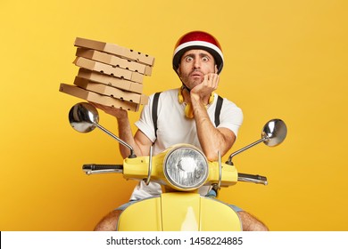 Photo of male courier has fearful expression, holds pile of cardboard boxes filled with pizza, delivers fast food for customers wears helmet poses over yellow background. Punctual delivery man on bike