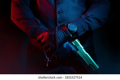 Photo of a male mafia criminal killer in suit and leather gloves holding a gun with cross on black background.