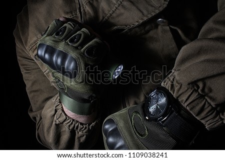 Photo of a male person in brown tactical outfit jacket and gloves using green tactical led flashlight and military watch.