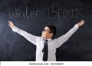 Photo Of A Male Primary School Student Standing In The Classroom With Text Of Winter Break On The Blackboard