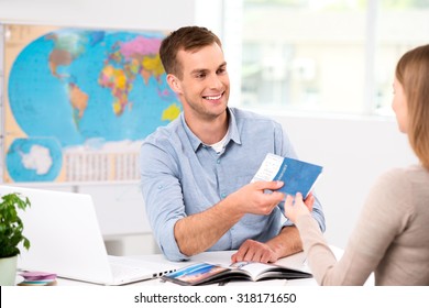 Photo of male travel agent and young woman. Young man smiling and giving tickets, passport with visa to female tourist. Travel agency office interior with big world map