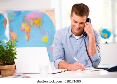 Photo of male travel agent. Young male operator smiling, talking on phone and offering vacation options for client. Travel agency office interior with big world map