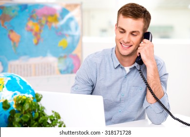 Photo Of Male Travel Agent. Young Man Talking On Phone And Smiling. Travel Agency Office Interior With Big World Map