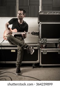 Photo of a man in his late 20's sitting in a rehearsal studio practicing his guitar playing.