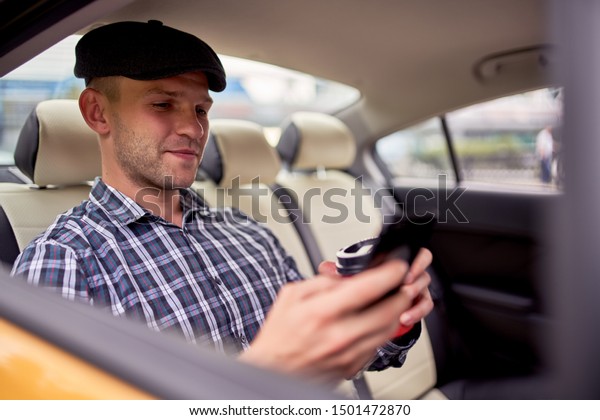 Photo of man with phone and glass of coffee sitting\
in back seat in taxi