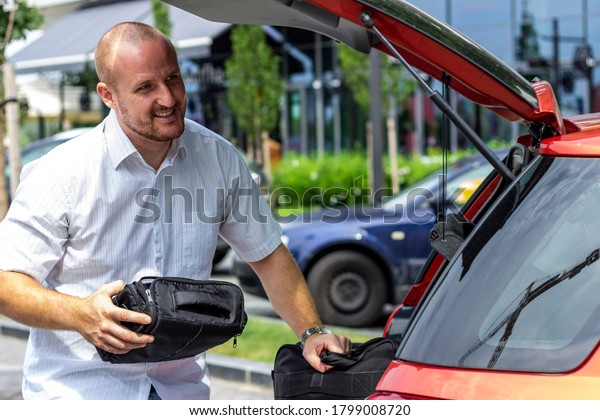 Photo of man putting bags in the car trunk.\
Businessman is unpacking the car trunk.Man packing luggage in car\
ready for road trip.Young Caucasian man loading the trunk of the\
car parked on the parking