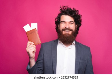A photo of a man smiling at the camera holding a passport with two tickets in it near a pink wall