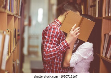 Photo Of Man And Woman Kissing In Library And Close Their Face By Book. Young Couple Of Boy And Girl Hiding For Spending Time Together. Students