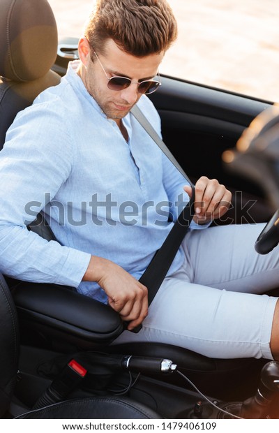 Photo of masculine\
man in sunglasses wearing seat belt while driving convertible car\
by seaside at sunrise
