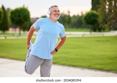 Photo Of Mature Active Positive Man In Wireless Headphones Doing Legs Stretching Exercises With Smile, Warming Up Muscles Before Routine Workout And Jogging Outside In City Park In Sunny Morning