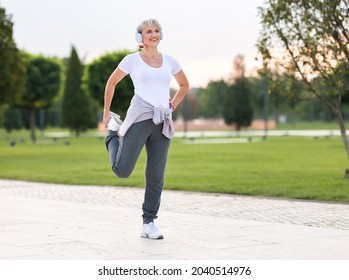 Photo of mature active positive woman in wireless headphones doing legs stretching exercises with smile, warming up muscles before routine workout and jogging outside in city park in sunny morning - Powered by Shutterstock