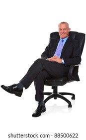 Photo Of A Mature Businessman Wearing A Smart Suit And Tie, Sat In A Leather Executive Chair With His Legs Crossed And Smiling To Camera, Isolated On A White Background With Natural Chair Reflection.