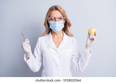 Photo Of Mature Dentis Woman In Facemask And White Coat Hold Stomatology Equipment And Apple Isolated On Grey Color Background