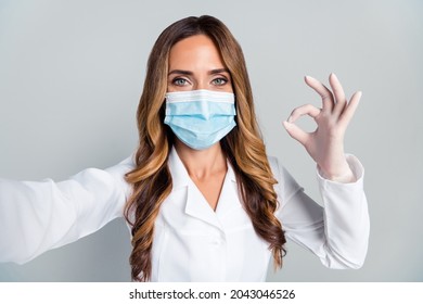 Photo Of Mature Happy Woman Hold Camera Make Selfie Okay Sign Doctor Wear Mask Isolated On Grey Color Background