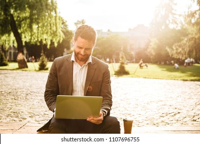 Photo Of Mature Joyous Man In Gray Classical Suit Sitting In Park With Takeaway Coffee While Working On Silver Laptop