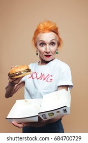 Photo Of Mature Redhead Fashion Woman Standing Isolated At Studio Eating Burger. Looking Camera.