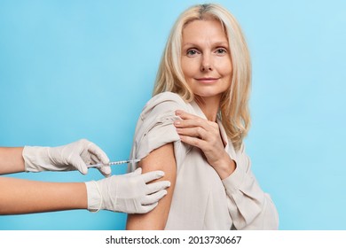 Photo Of Middle Aged Woman With Fair Hair Gets Injection As Protecton Against Coronavirus During Quarantine Looks Self Confident At Camera. Unknown Doctor Gives Vaccinationn Shot To Patient.