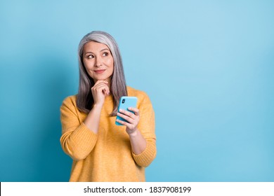 Photo of minded old woman use smartphone look copyspace touch fingers chin think thoughts decide texting typing chatting wear style stylish sweater isolated blue color background