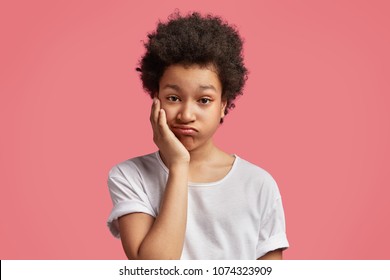 Photo Of Mixed Race Small Kid Feels Bored, Wants To Play With Parent Who Are Busy, Has Crisp Bushy Hairstyle, Dressed Casually. African American Schoolboy Has Toothache, Keeps Hand On Cheek Feels Pain