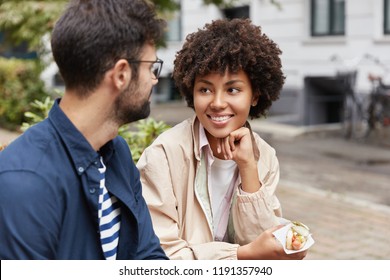 Photo of mixed race tourist enjoy lively communication, discuss traditions of their countries, eat fastfood on street, being in good mood. Beautiful Afro American girl talks with handsome guy outdoor - Shutterstock ID 1191357940