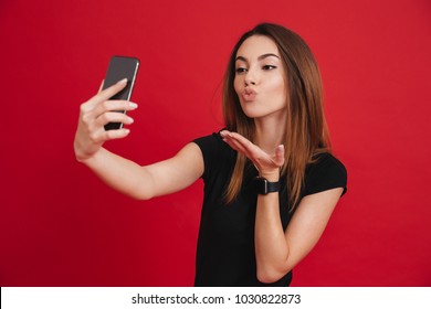 Photo of modern woman in black t-shirt partying and making air kiss on mobile phone, isolated over red background