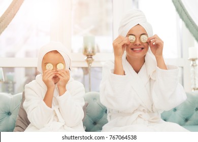 Photo of mother and daughter in white bathrobes. They are sitting on the couch and applying pieces of cucumber to their eyes. Their hair is wrapped in a white bath towel. - Powered by Shutterstock