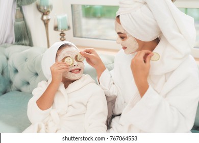 Photo of mother and daughter in white bathrobes. They are sitting on the couch and applying pieces of cucumber to their eyes. Their hair is wrapped in a white bath towel. They have cucumber mask. - Powered by Shutterstock