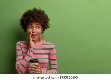Photo of mysterious woman with curly hair, keeps palm near mouth, tells secret, gossips with someone while drinking coffee, looks suspiciously aside, wears striped jumper, copy space on green