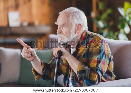 Photo of nervous angry retired man dressed plaid shirt holding nursery cane pointing finger sitting couch indoors apartment
