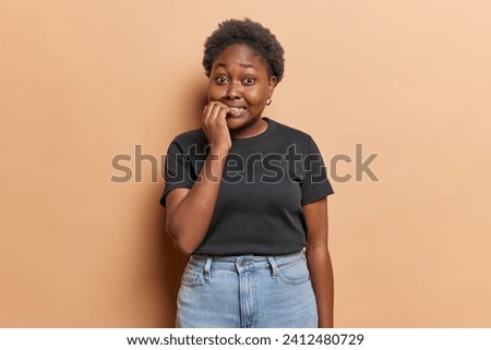 Photo of nervous overweight dark skinned young woman bites finger nails looks anxious at camera dressed in casual black t shirt and jeans isolated over brown background feels stressed or worried