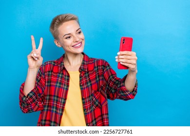 Photo of nice girl with short hairstyle dressed red shirt holding smartphone doing selfie show v-sign isolated on blue color background