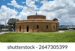 Photo of an old circular building, former ammunition depot, currently a Popular Theater in the middle of the square surrounded by green grass, blue sky with many white clouds, beautiful landscape