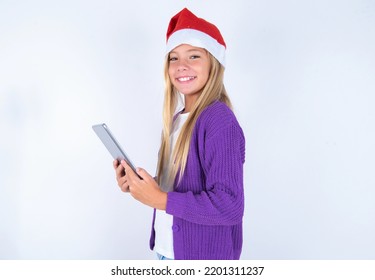 Photo Of Optimistic Little Kid Girl With Christmas Hat Wearing Yarn Jacket Over White Background Hold Tablet