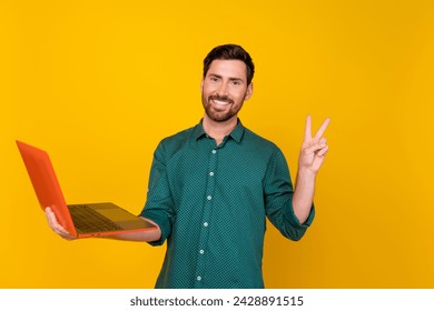Photo of optimistic nice guy with beard dressed dotted shirt hold laptop showing v-sign symbol isolated on vivid yellow color background