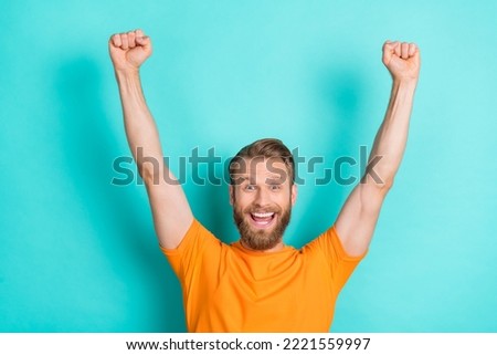 Photo of optimistic satisfied crazy man with blond hairstyle dressed orange t-shirt raise fists up isolated on teal color background