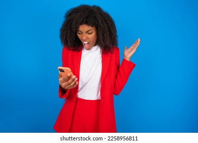 Photo of outraged annoyed young businesswoman with afro hairstyle wearing red over blue wall holds cell phone, makes call, argues with colleague,  expresses negative emotions. People and anger. - Shutterstock ID 2258956811