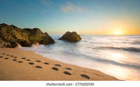 Photo Of The Pebble Beach Of Cowell Ranch Near Half Moon Bay On The California Coast.  Sunset Scene With Ocean Waves And Footprint Walking Across The Beach.