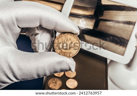 Photo of a person's male hand in white gloves holding a 1940 half penny british coin on stack of books and magnifyng glass background. Numismatics hobby concept.