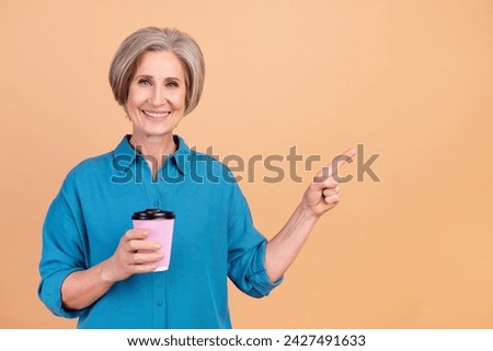Photo of pleasant woman dressed stylish shirt holding cup of cappuccino indicating at offer empty space isolated on beige color background