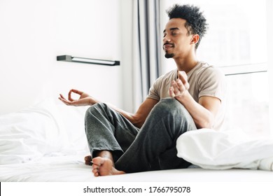 Photo of pleased african american man with mustache meditating while sitting on bed at bright room