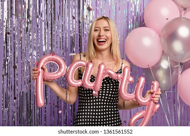 Photo of pleased blonde woman with make up, has some sort of shindig, holds balloons, poses over flying confetti, tinsel, being on night out, hangs out on party with friends during Friday night - Shutterstock ID 1361561552