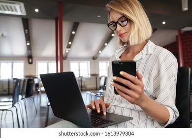 Photo of pleased blonde woman wearing eyeglasses using cellphone and laptop while sitting in conference hall