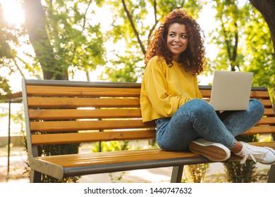 Photo of a pleased cheerful young beautiful curly student girl sitting outdoors in nature park using laptop computer.