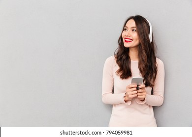 Photo of pleased female smiling looking aside with silver smartphone in hands enjoying favorite music, via wireless headphones over gray background