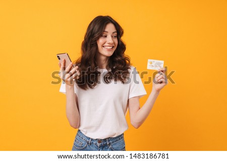 Photo of pleased young woman posing isolated over yellow wall background using mobile phone holding debit card.