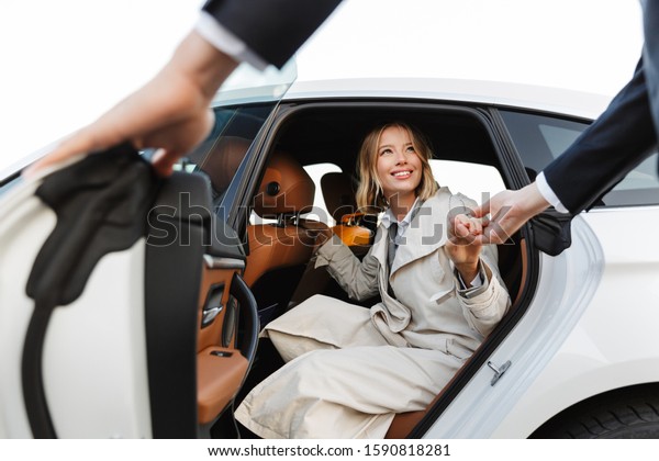 Photo of polite man in formal\
wear helping nice businesswoman to get out of car on parking\
outdoors