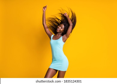 Photo portrait of african american girl dancing with both hand in the air hair flying, wearing blue dress isolated on bright yellow colored background